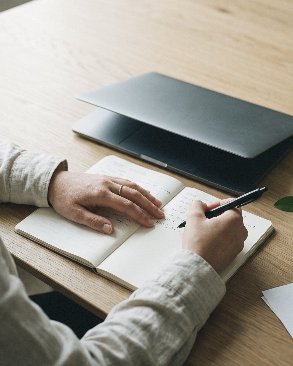 Manos escribiendo a mano en un cuaderno abierto, junto a un portátil cerrado sobre una mesa de madera.